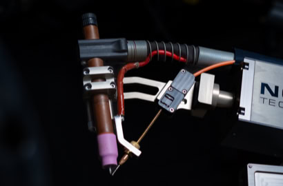 Close-up of a machine part with a metal arm and a brown cylindrical component, featuring red and orange wires.