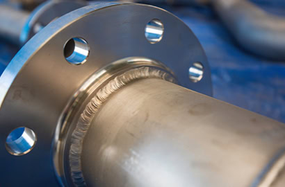 Close-up of a metal pipe with a flange and visible welding seam. The pipe has several bolt holes around the flange edge.