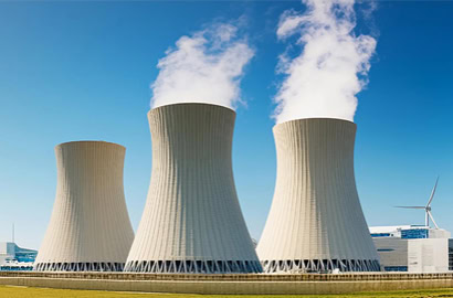 Three large cooling towers emit steam against a clear blue sky. A wind turbine is visible in the background to the right, surrounded by an industrial setting with green grass in the foreground.