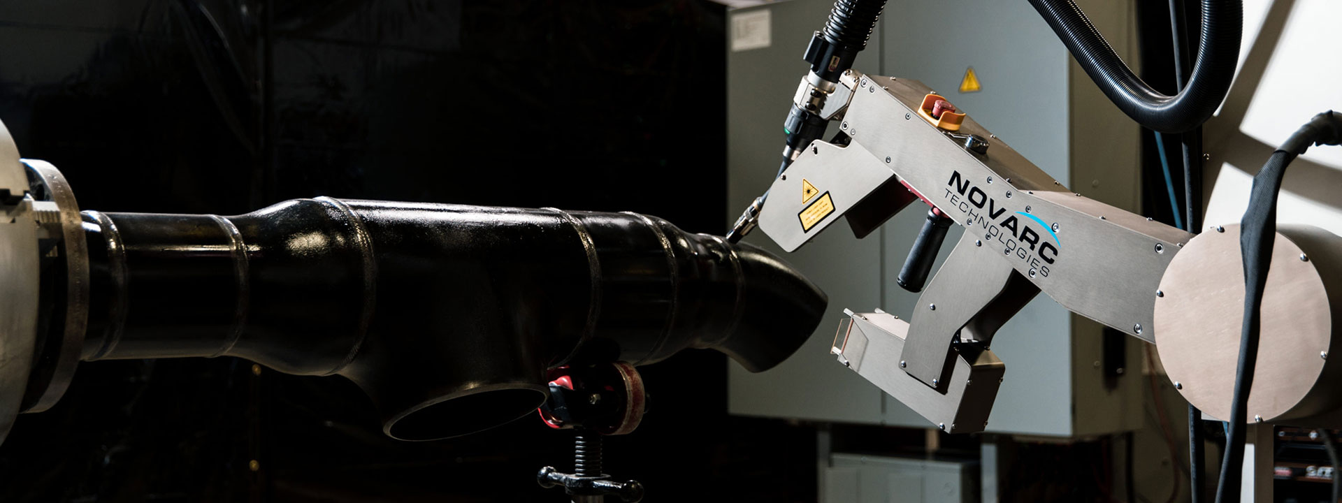 A robotic welding arm labeled "Novarc" is working on a large black metal pipe fitting in an industrial setting.