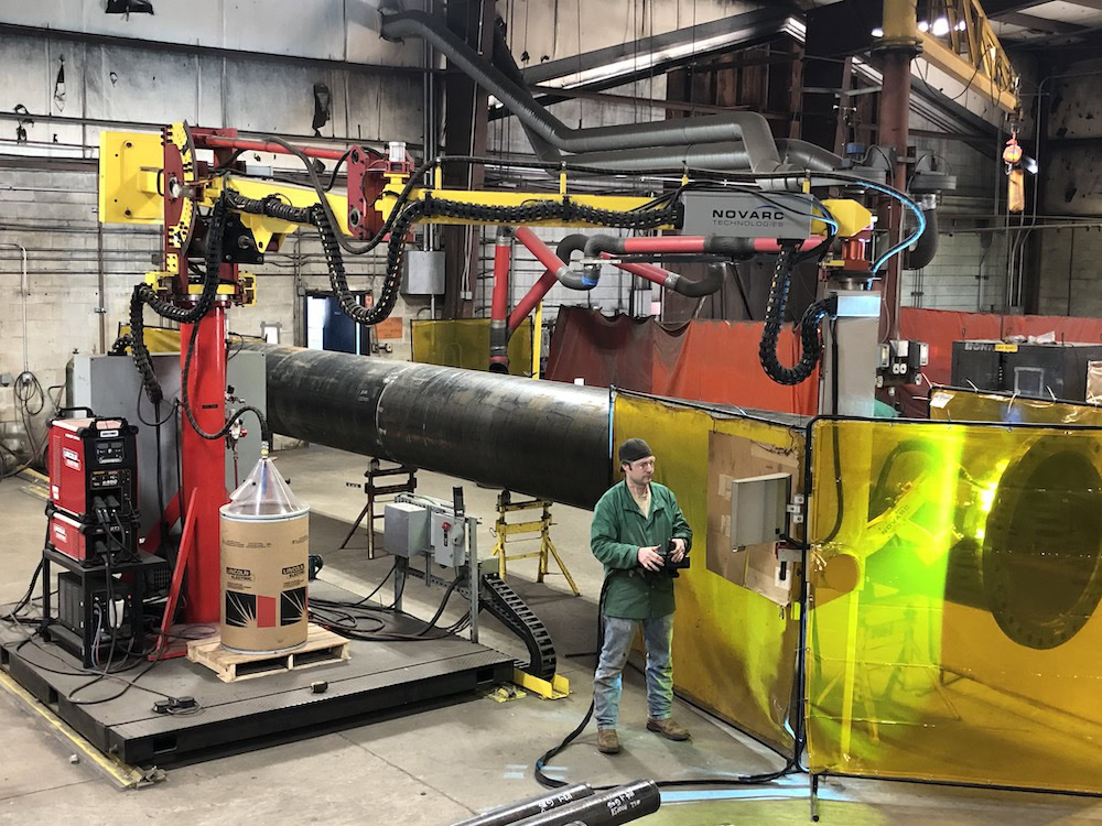 A worker operates a large industrial machine in a factory setting. The machinery involves a long cylindrical pipe and is partially enclosed by yellow safety screens. The factory environment appears spacious with various equipment and cables.