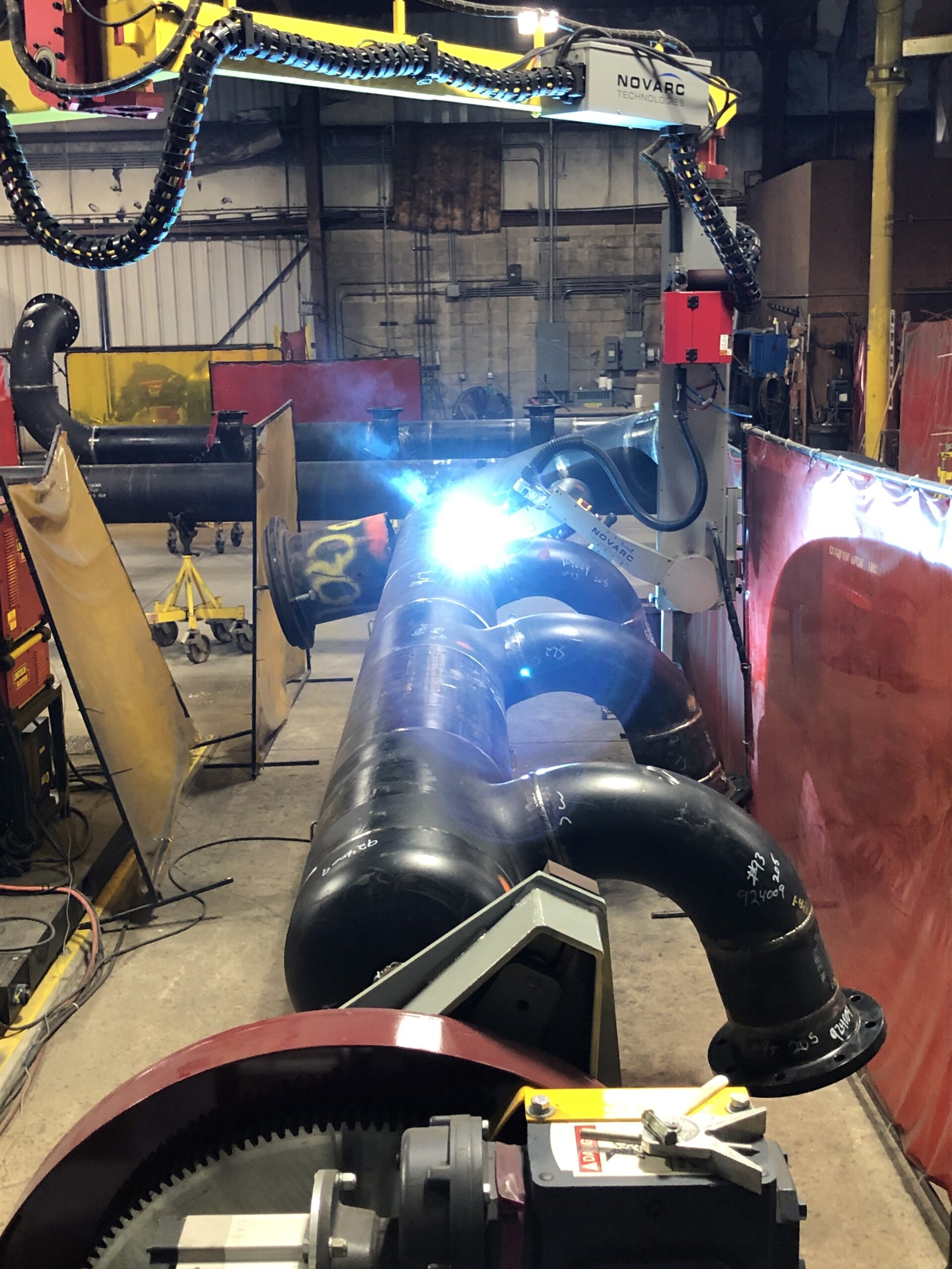 A workshop featuring industrial welding equipment and large metal pipes being welded. Bright welding sparks are visible as robotic arms and protective screens surround the area, with various machinery and safety barriers in the background.