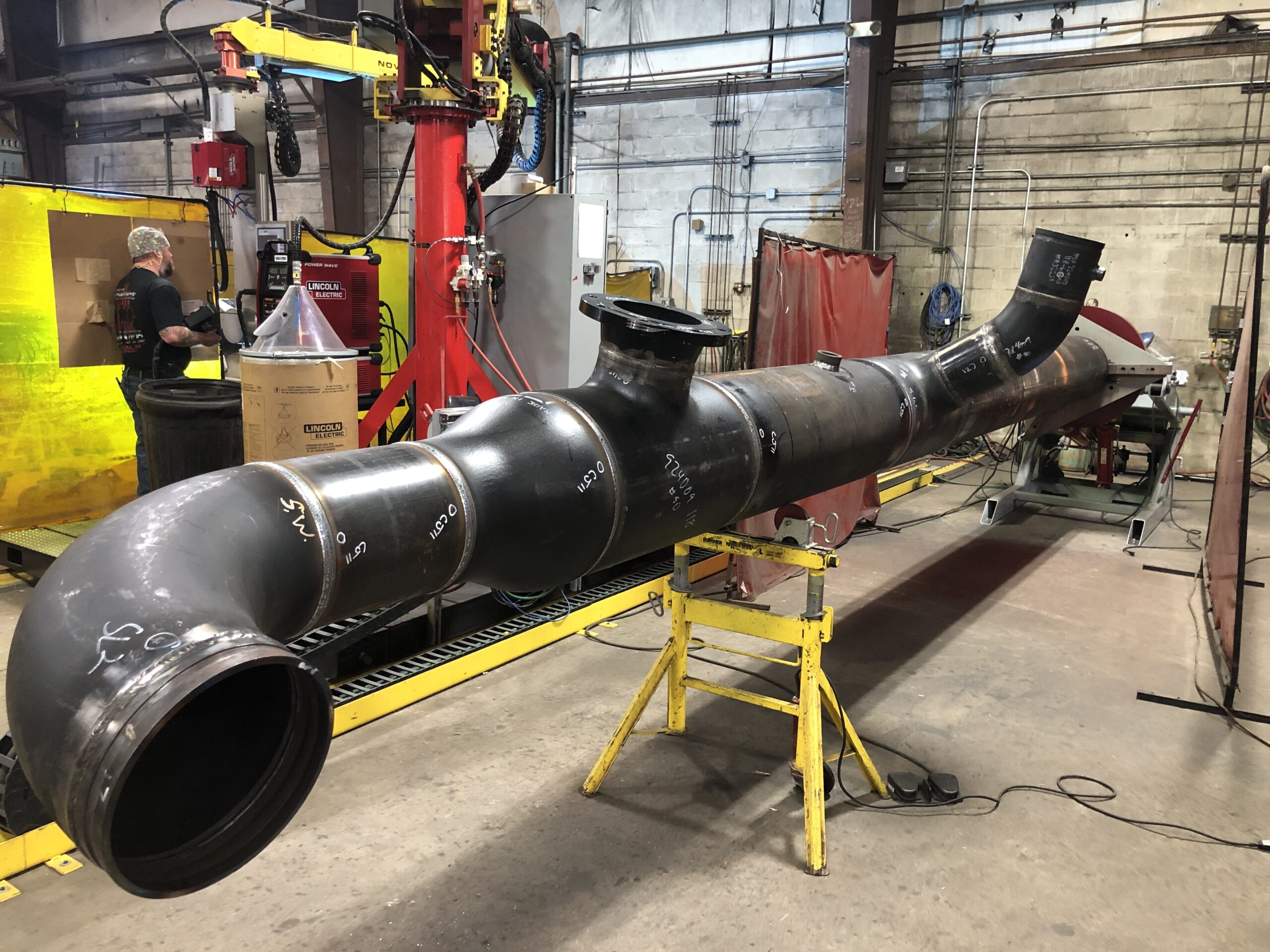 A long, black metal pipe with various bends and openings is set on yellow supports in an industrial workshop. Workers and machinery are visible in the background, along with protective curtains and equipment.