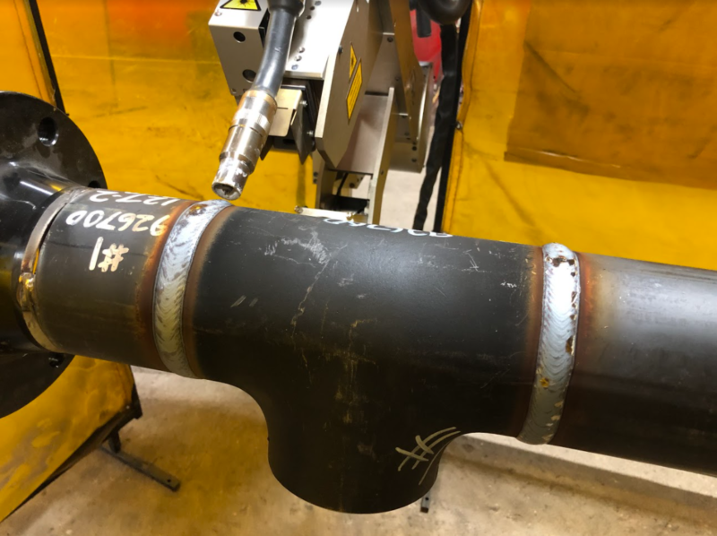 A welding machine is positioned to weld a dark metal pipe with visible markings and seams. The surrounding area includes a yellow protective screen and concrete floor.