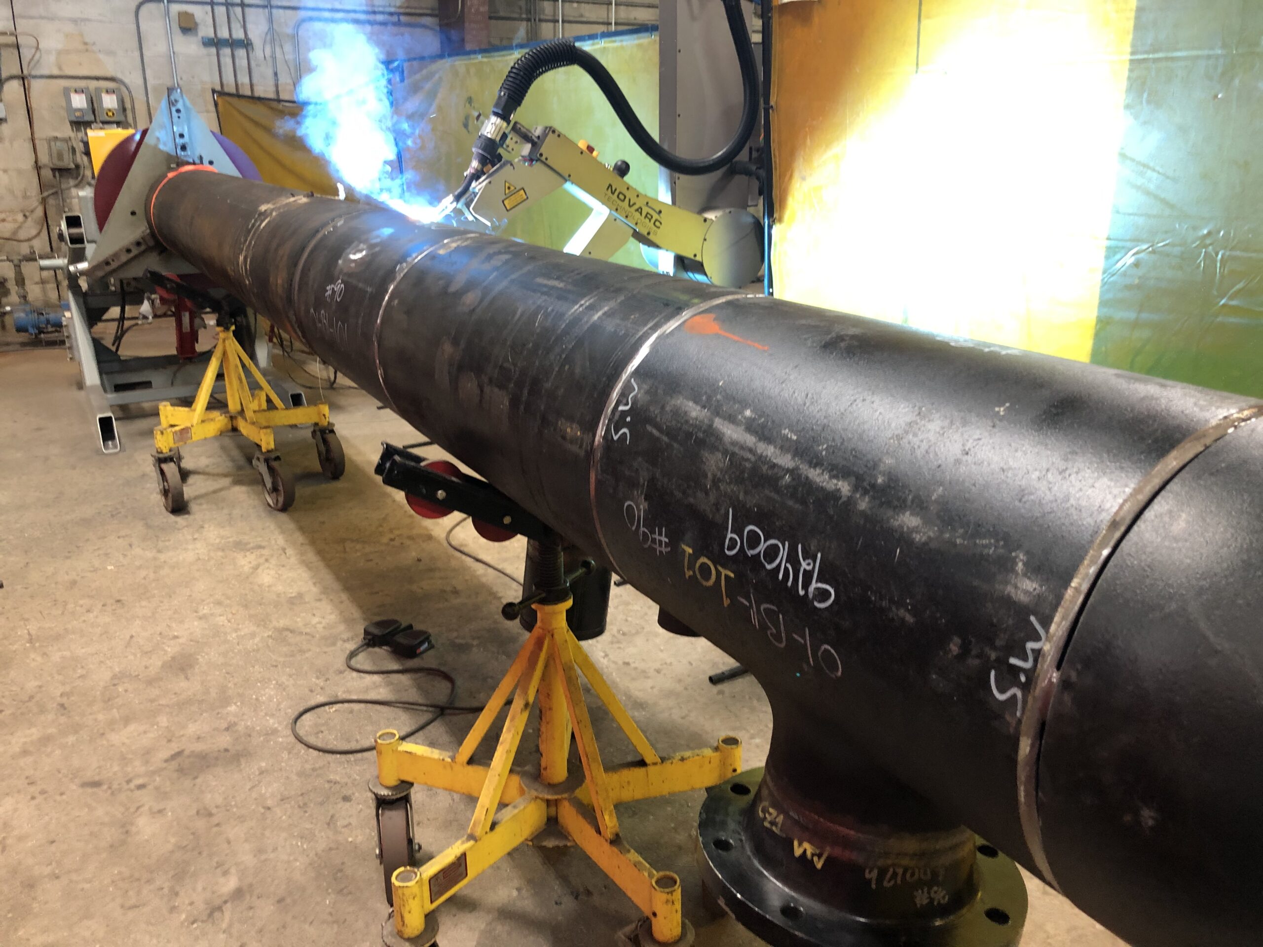 A large metal pipe is secured on adjustable stands in an industrial setting. A robotic arm welds the pipe, emitting sparks and blue light. The background features yellow and gray walls, with various equipment visible.