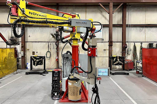 A robotic welding arm in a workshop. The yellow arm labeled "Novarc" is mounted on a red base, surrounded by various welding equipment. A control panel with a digital screen is attached to the setup, and the space is organized with metal walls.