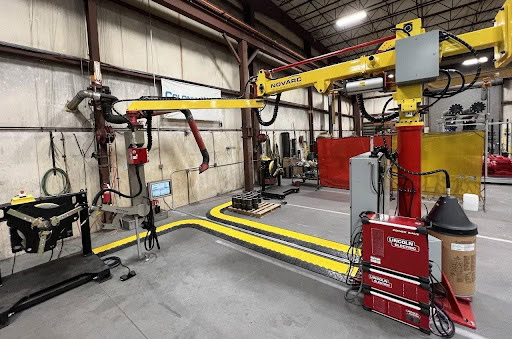 A workshop features an automated welding station with robotic arms and welding equipment. There are machines labeled "Novarc" and "Lincoln Electric" and components on pallets. The area is marked with yellow safety tape.