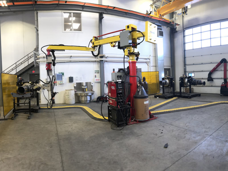 A spacious industrial workshop with welding equipment and a robotic arm. The area has safety barriers and a large garage door to the right. Various metalwork tools and machinery are spread throughout the well-lit space.