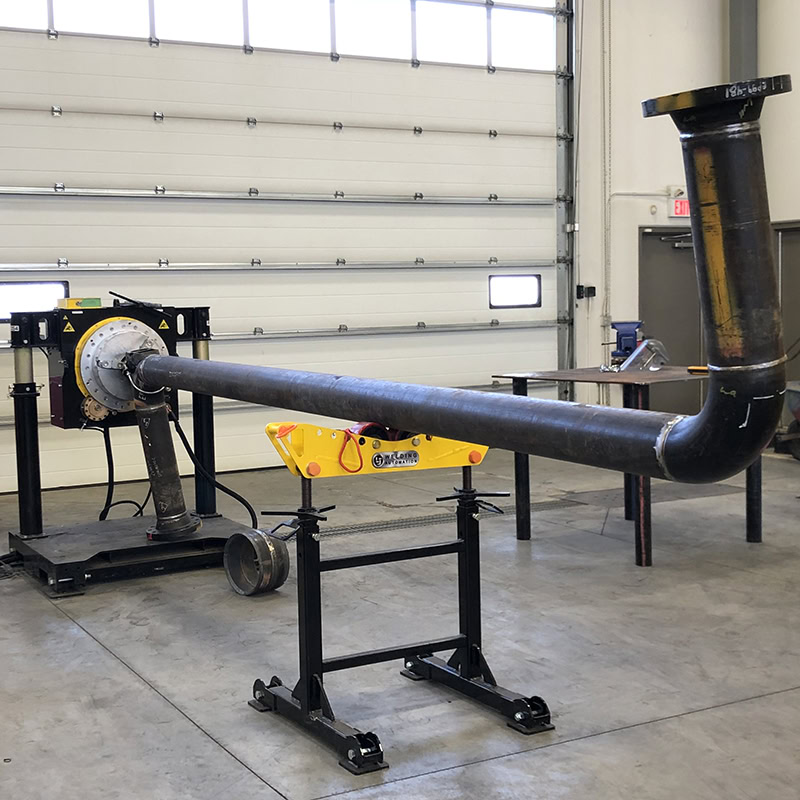 A large metal pipe is set up horizontally on two supports inside a workshop. The pipe has a 90-degree bend at one end and is connected to machinery on the left. The workshop has a large garage door in the background.