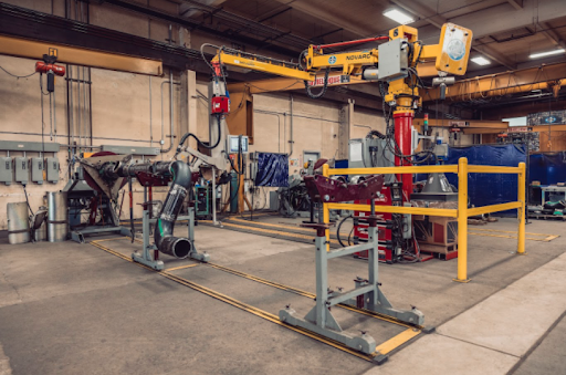 A large industrial robot arm is positioned in a spacious factory setting. Surrounding the arm are yellow safety barriers, various machinery, and tools. The walls are lined with panels and control boxes, and the floor is marked with safety lines.