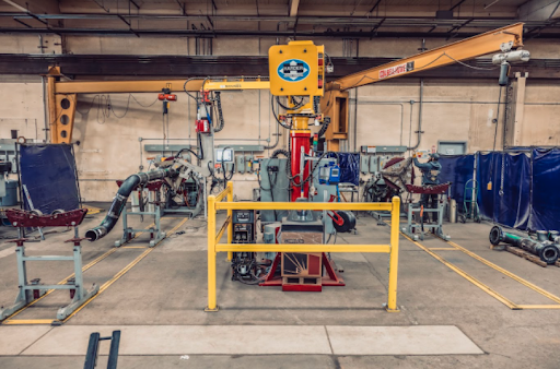 A factory workshop with a large yellow industrial machine and robotic arms surrounded by protective barriers. Various equipment and tools are scattered around the concrete floor, beneath high ceilings with exposed beams.