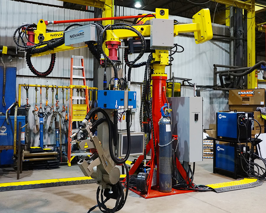 A robotic welding arm with various mechanical components and control panels in an industrial setting. Equipment surrounds the arm, including storage racks and machinery, under a high ceiling with exposed beams.