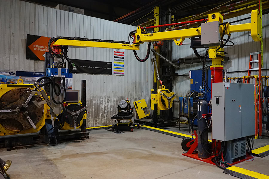 A manufacturing facility with a large yellow robotic arm connected to industrial machinery. The area is surrounded by cables, control panels, and other equipment, suggesting an automated process environment. Signs and safety markings are visible.