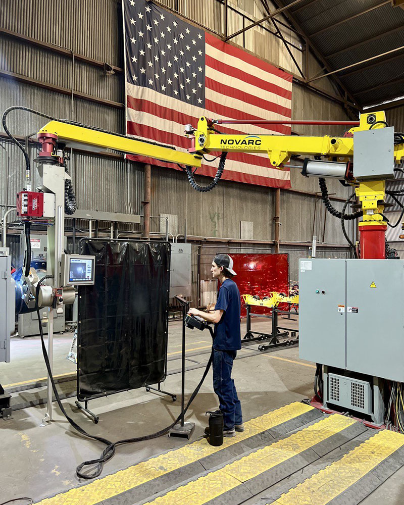 A person operates a welding robot in an industrial setting. The American flag hangs in the background. The workshop is equipped with metal equipment and safety barriers. The person is focused on controlling the machinery.