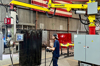 A person operates a robotic welding machine in an industrial setting. The robot arm, labeled "Novarc," is suspended from above. The workspace has various equipment and safety barriers, with an American flag in the background.