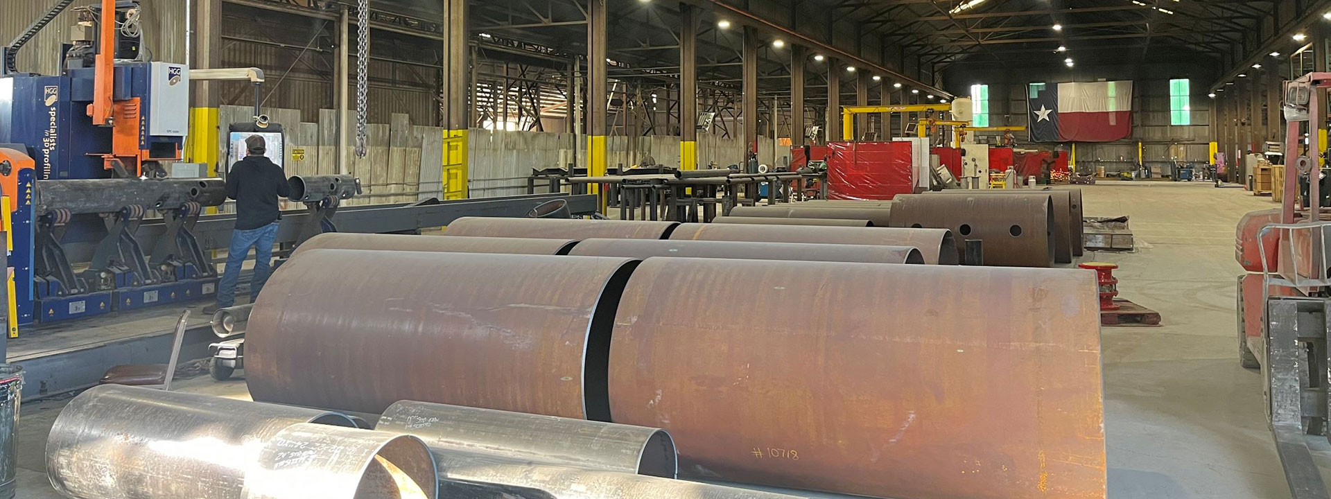 A large industrial workshop with several large metal pipes on the floor. A person operates machinery on the left side.