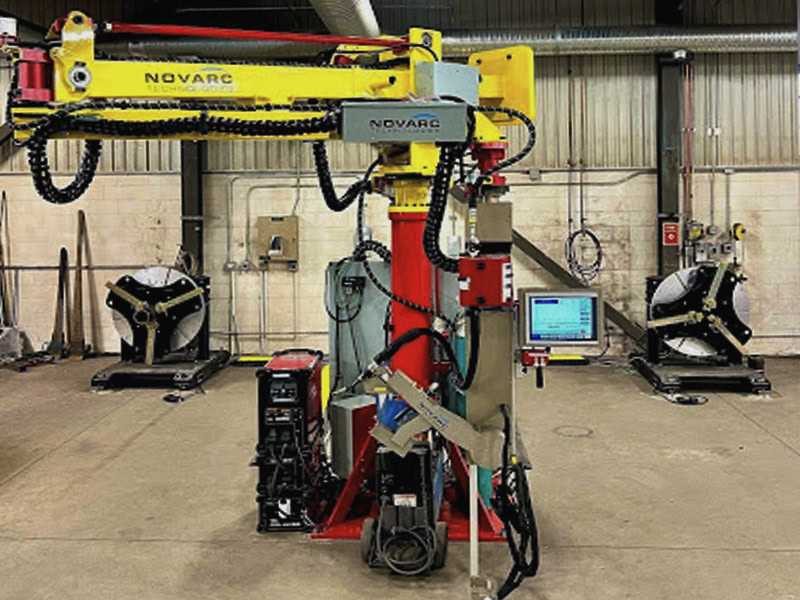 A large industrial welding robot stands in a workshop. It features a yellow arm with "Novarc" branding and various cables attached. In the background, there are two workstations and a control panel display.