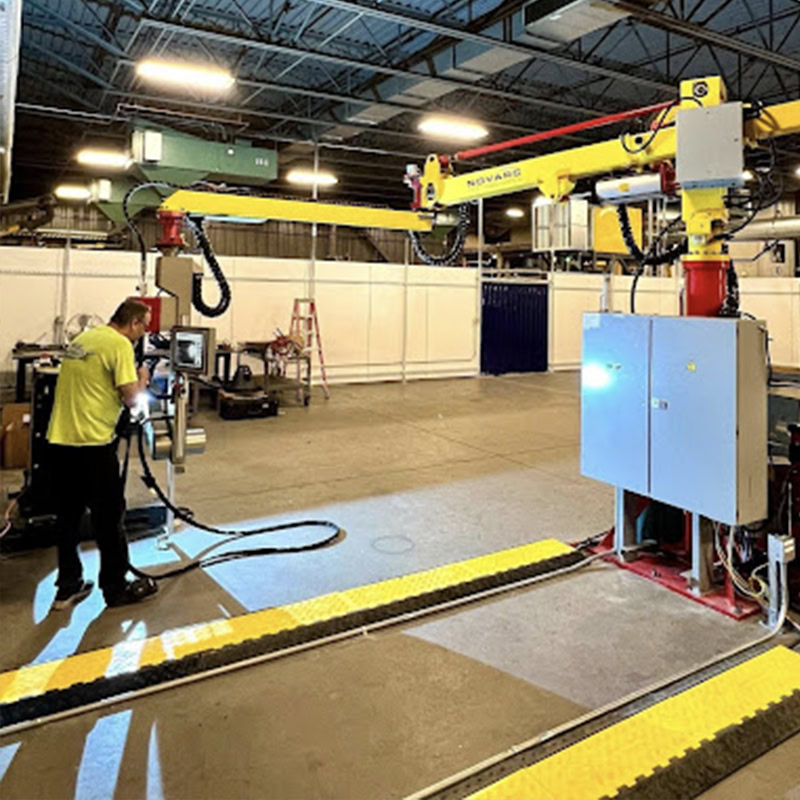 A worker in a high-visibility shirt operates machinery inside a large industrial facility. Overhead robotic arms are visible, and bright lights illuminate the space. A ladder and various equipment are in the background.