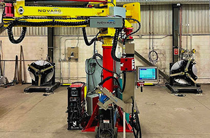 A robotic welding machine in a workshop. The machine is yellow and red, with monitors and cables attached. The industrial setting includes metal walls and additional equipment in the background.