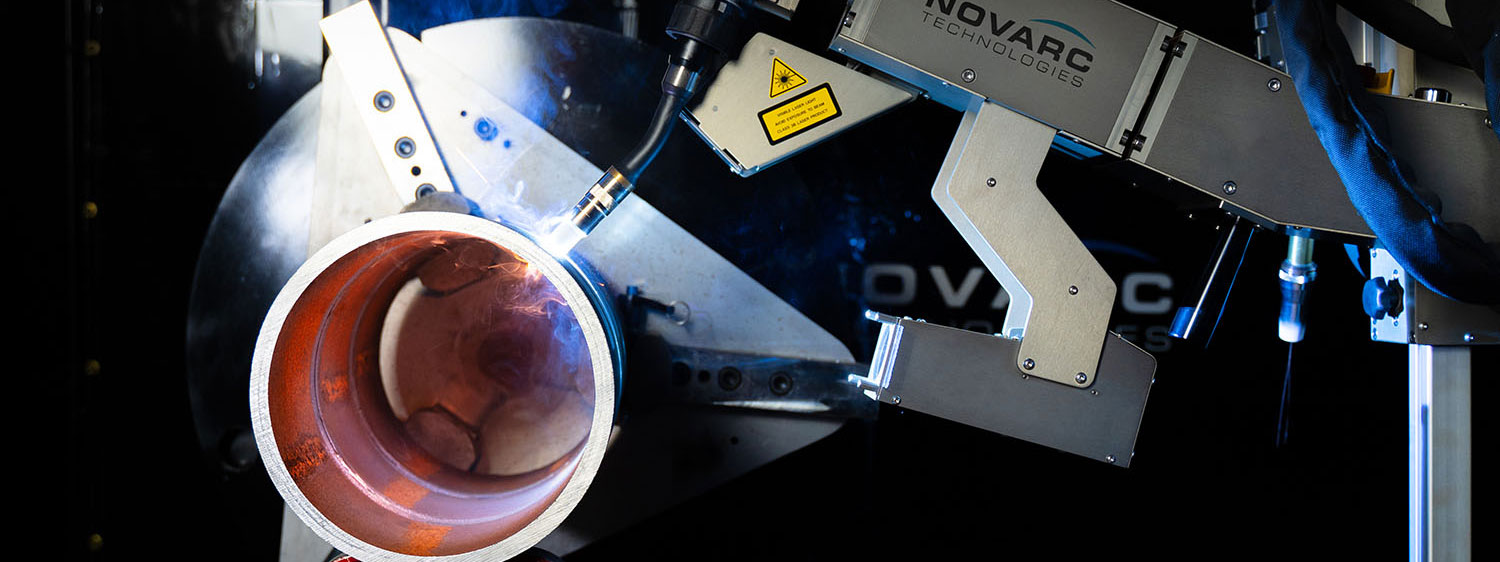 A robotic arm from Novarc Technologies welds a large metal pipe. The machine is illuminated against a dark background.