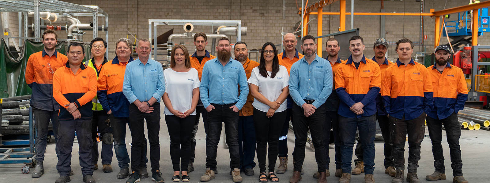 A group of people stands in a workshop setting. Some wear orange and navy work uniforms, while others are in casual light blue and white shirts. The background features industrial equipment and piping.