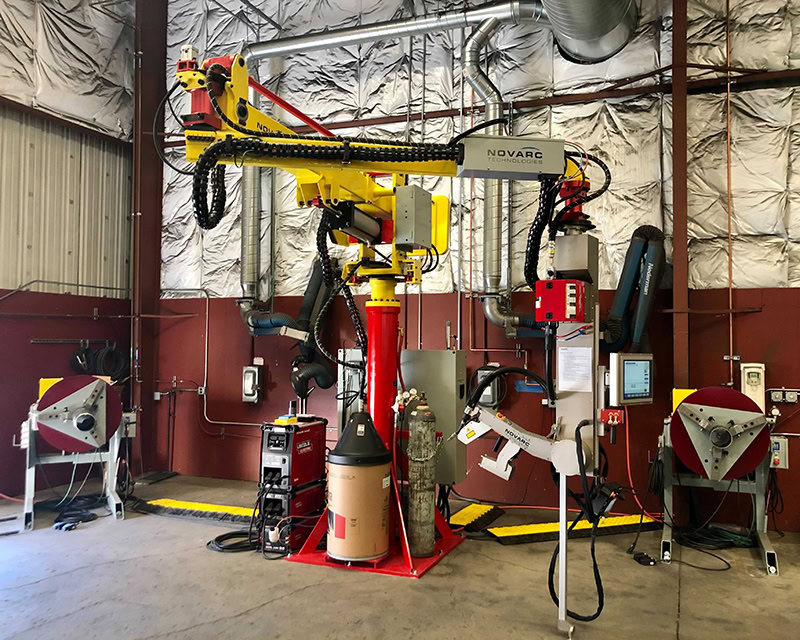 A large industrial robotic welding machine with a central arm and attachments, surrounded by equipment and control panels, is situated in a metal-walled workshop. The setup is part of a mechanized welding system.