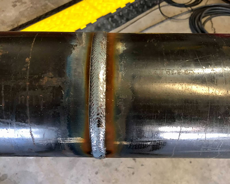 Close-up of a welded metal pipe section with noticeable weld bead along the joint. The metallic surface shows signs of heat discoloration around the weld, with a dark sheen and surrounding equipment in the background.