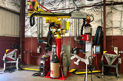 A welding robot in a large industrial space. The robotic arm is yellow and red with cables and machinery surrounding it. The walls have metallic insulation, and various control panels and equipment are nearby.