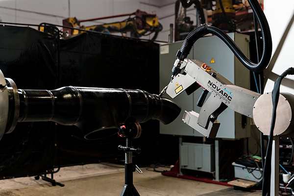 A robotic arm equipped with welding tools operates in a manufacturing setting. It is positioned to work on a metal component. The background shows machinery and industrial equipment, with curtains partially enclosing the work area.