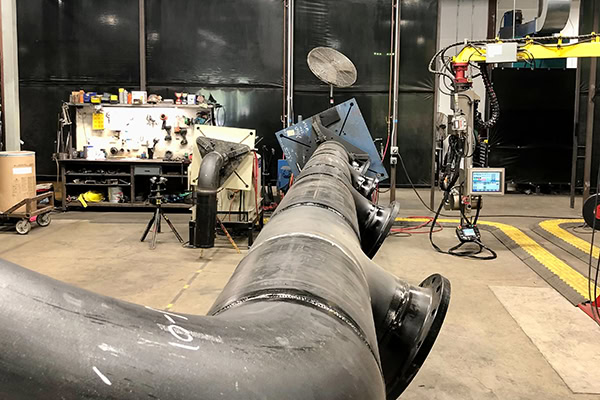 A large industrial pipe setup in a workshop. Various tools and equipment are visible, along with monitors and machinery. Workbenches with assorted items are in the background, contributing to a busy, functional environment.