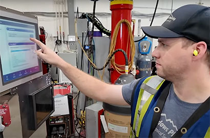 A worker wearing a cap, safety vest, and earplugs operates a touchscreen control panel in an industrial setting. Various equipment, wires, and a fire extinguisher are visible in the background.