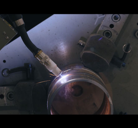 Close-up of a welding process on a metal cylinder, with a welding torch emitting sparks. The setup includes clamps and tools holding the cylinder in place