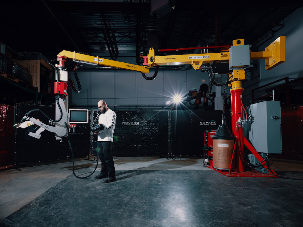 A person operates a large robotic welding arm in an industrial setting. The area is equipped with safety barriers and overhead lighting. The robotic arm is attached to a red and yellow support and control panel.