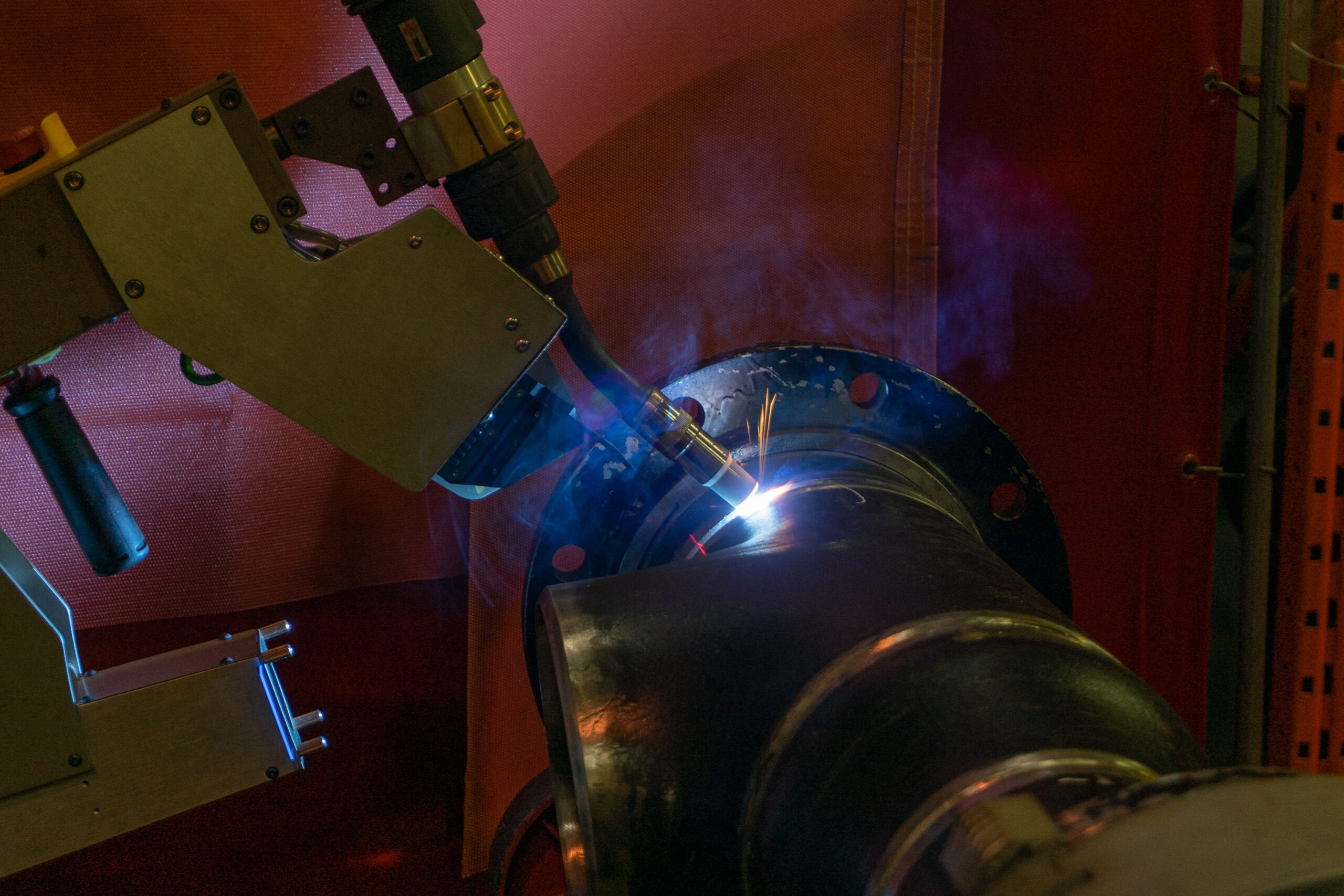 A robotic welding machine is working on a large metal pipe, emitting bright sparks and smoke. The setup is inside a workshop against a red backdrop. The machinery is positioned at the pipe's joint, performing automated welding.