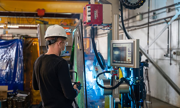 A person wearing a hard hat and mask operates machinery in an industrial setting. They are interacting with a touchscreen display, surrounded by various equipment and safety barriers.