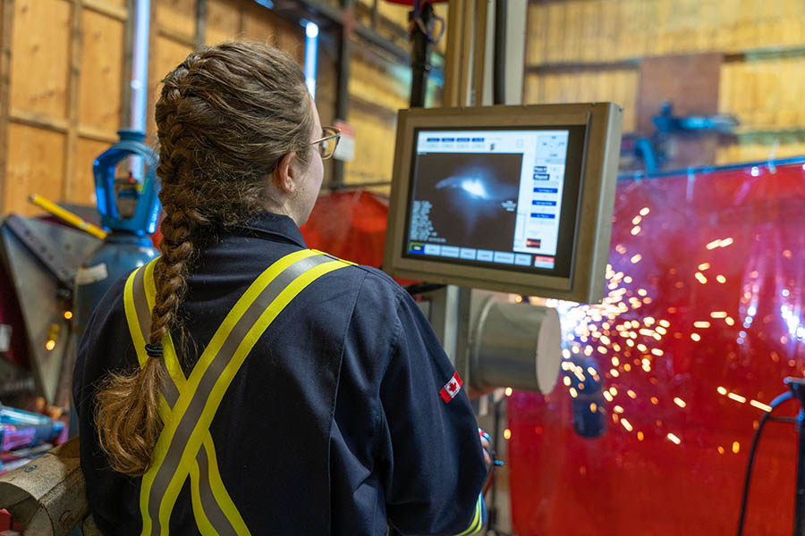 A person in a reflective safety uniform monitors a welding operation on a computer screen. Sparks from welding are visible in the background, and a Canadian flag patch is on the sleeve of the uniform.