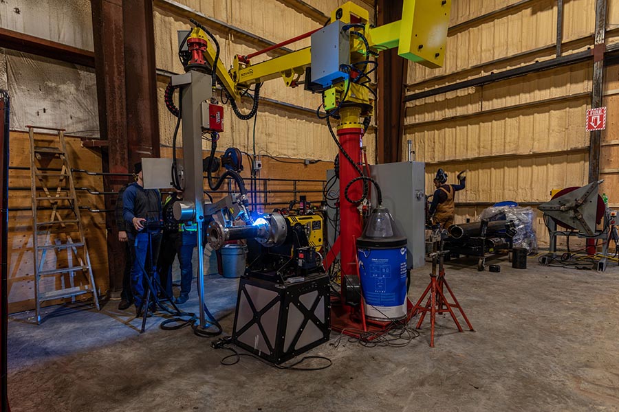 A workshop scene with machinery and equipment. Two people operate a large industrial welding machine, surrounded by various tools, pipes, and a ladder. The space is enclosed with unfinished wooden walls and a concrete floor.