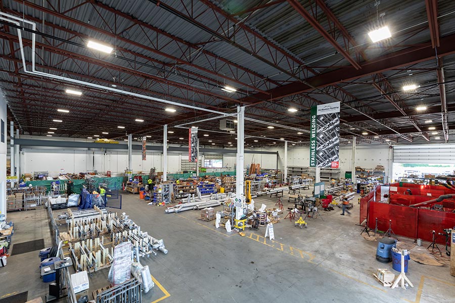 A spacious industrial warehouse with high ceilings, containing various machinery and equipment. Workers are scattered throughout, engaging in different tasks. Shelving and materials are neatly organized along the walls. Ample lighting illuminates the area.