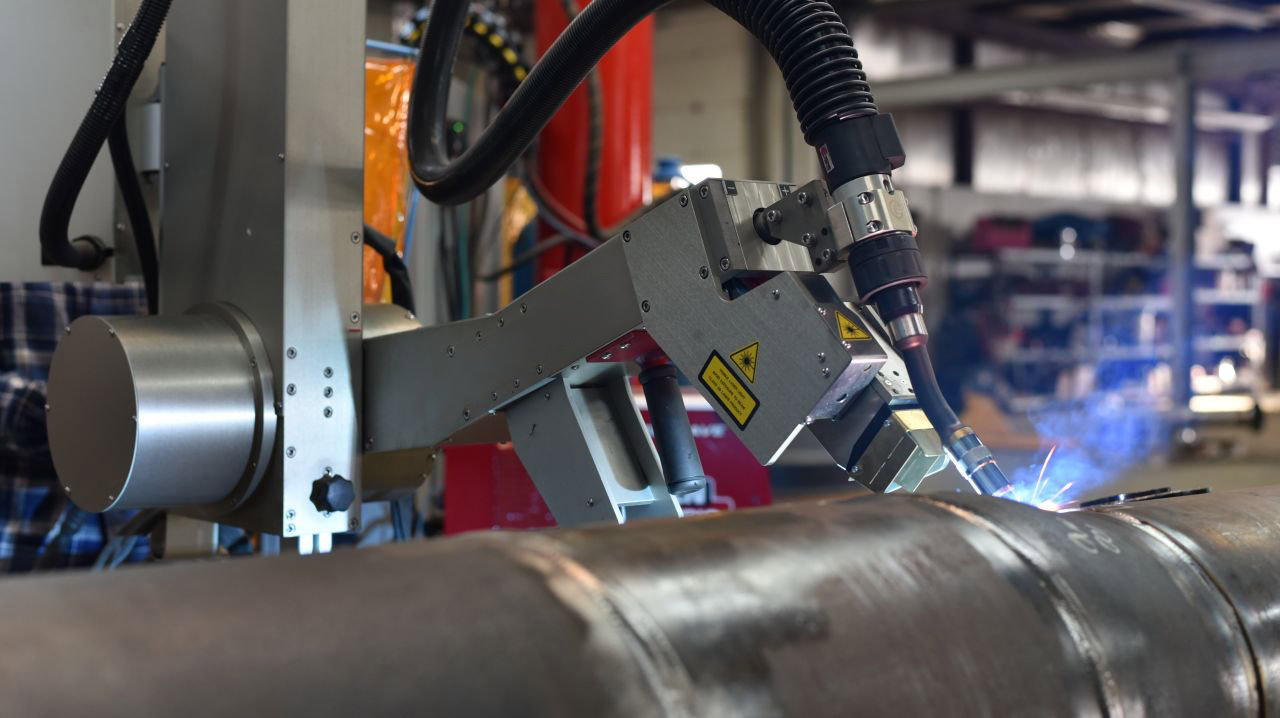 A robotic arm equipped with a welding torch is performing welding on a metal pipe. Sparks are visible from the welding process. The background shows shelves with various equipment and tools in a workshop setting.