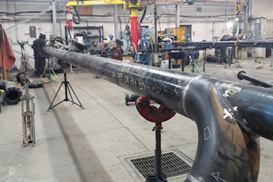 A large industrial pipe is being welded in a workshop. Several workers wearing protective gear are visible in the background. The workshop is equipped with tools, safety equipment, and machinery, while the pipe rests on a tripod stand.