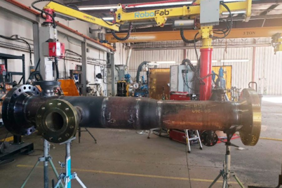 A large industrial pipe section is held up by supports in a factory setting. Overhead cranes and various equipment, including a "RoboFab" unit, are visible in the background, suggesting a manufacturing or assembly environment.