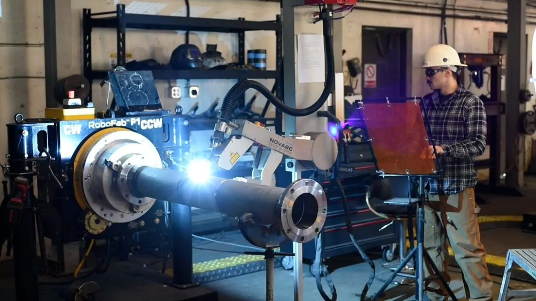 A worker in a helmet and goggles operates a large industrial machine with a robotic arm inside a workshop. The arm is working on a cylindrical metal component, with a bright light emanating from the process. Tools and equipment are visible in the background.