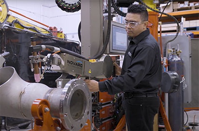 A technician operates a Novarc welding robot on a large metal pipe. He is adjusting the machine while wearing safety glasses, in an industrial setting with various equipment and tools in the background.