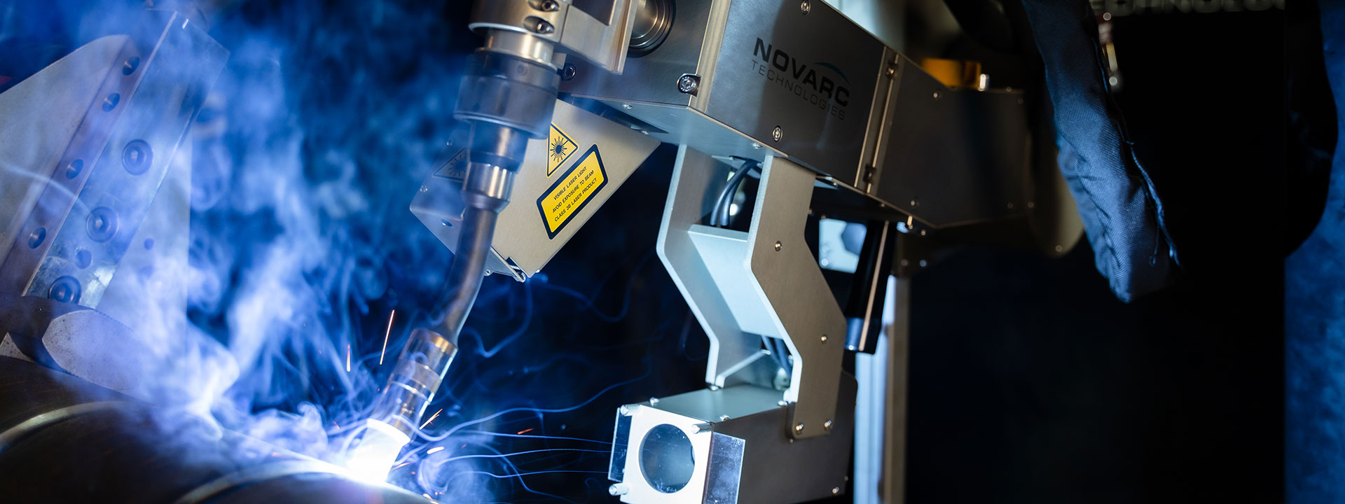 A close-up of an industrial welding robot in action, emitting bright sparks and blue light as it works on metal pipes. The machine features a metallic arm and visible wiring, with a brand label on its body. The background is slightly blurred.