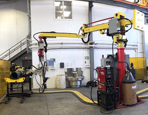 A large yellow and red industrial robotic arm is positioned in a spacious factory setting. The arm is equipped with various tools and connected to machinery