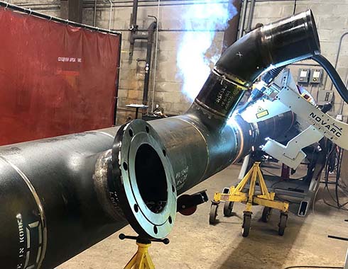 A welding robot operates on a large metal pipe in an industrial workshop, emitting bright sparks.