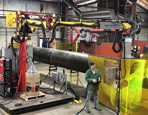 A worker stands next to industrial machinery in a factory. The equipment includes robotic arms and a large cylindrical object.