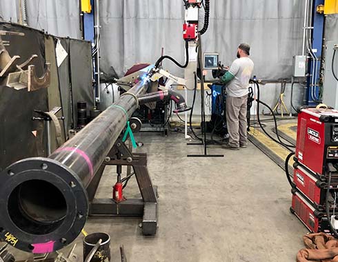 A man operates a machine in an industrial workshop. A long metal pipe is mounted on a stand. Various tools and equipment are scattered around, with a curtain in the background.