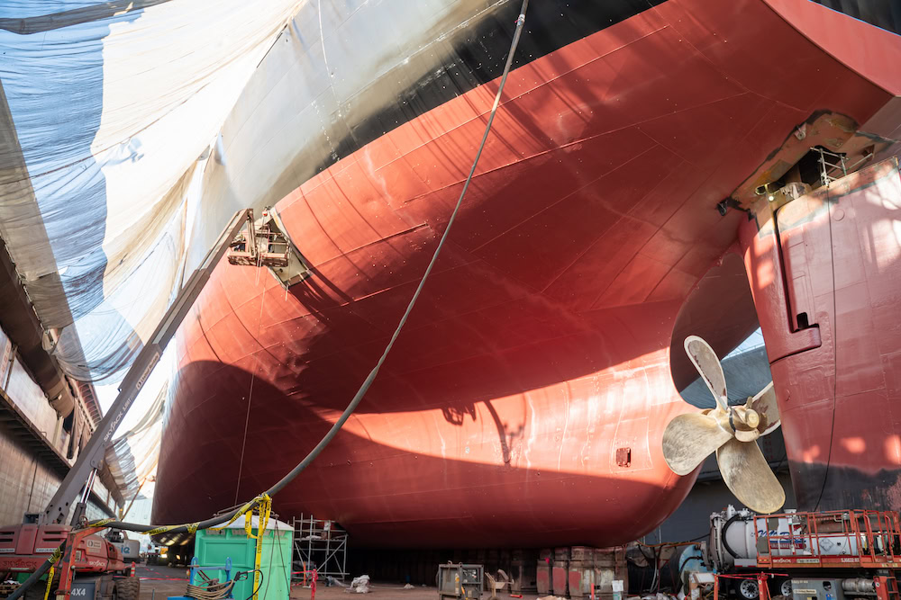 View of a large ship undergoing maintenance in a dry dock, with its red hull exposed. A propeller is visible on the right, alongside scaffolding and various equipment. White tarps partially cover the top sections.