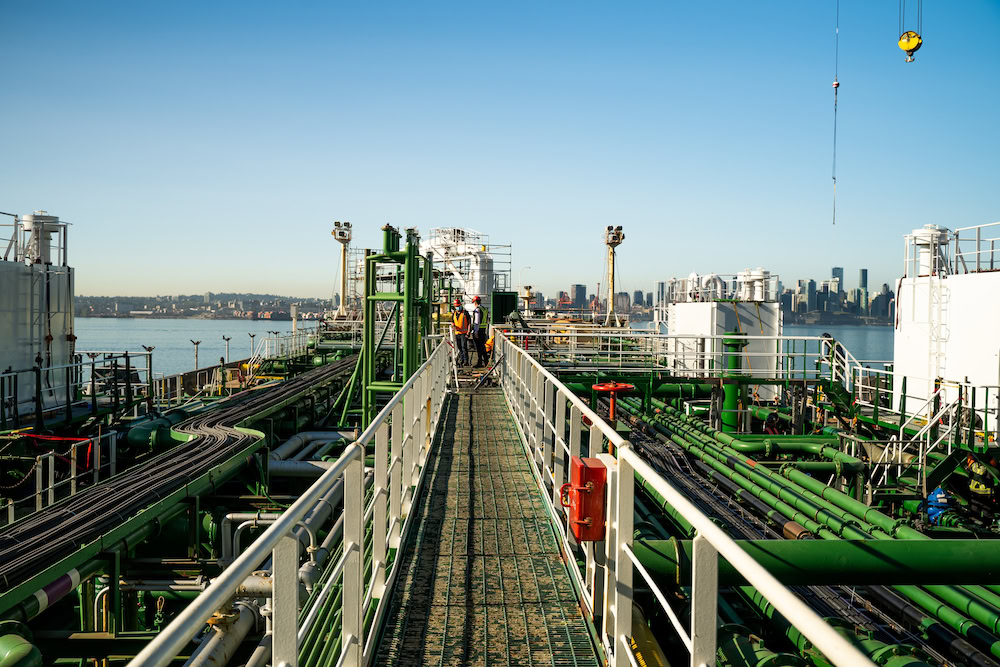 View from the deck of a large ship with green pipes and railings, facing a city skyline in the distance under a clear blue sky. An industrial setting with a crane visible on the right side.